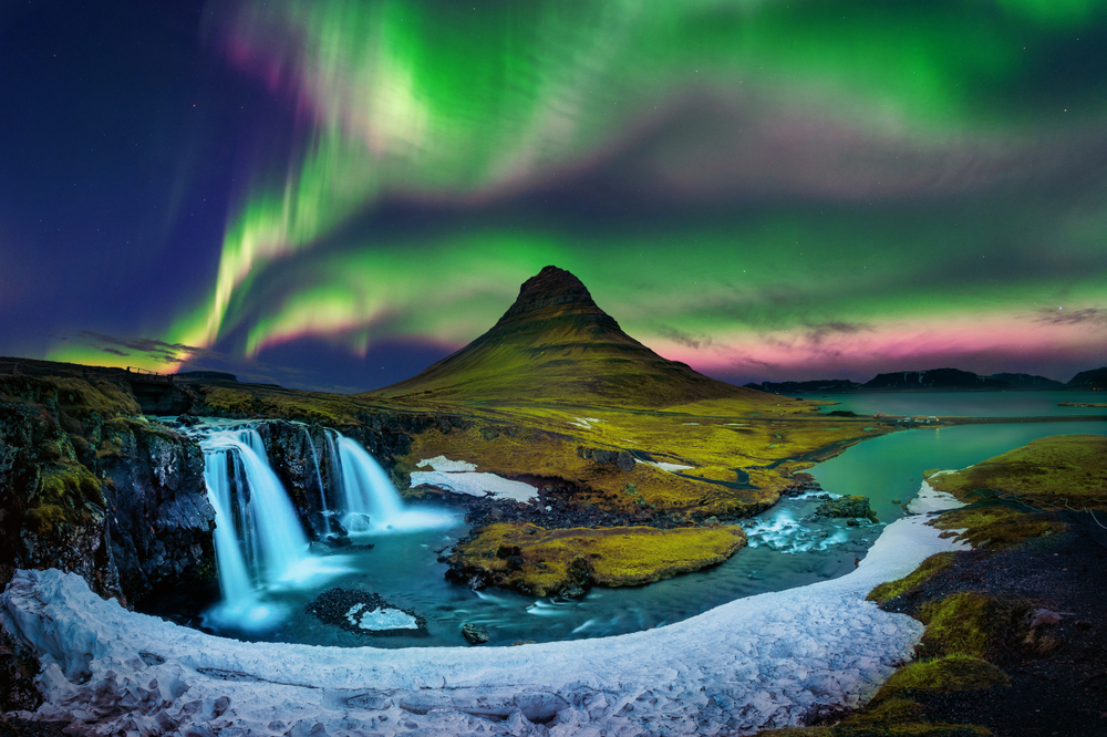 Aurora boreal verde y violeta sobre la montaña Kirkjufell y una cascada, con río y nieve en primer plano, en una noche clara.