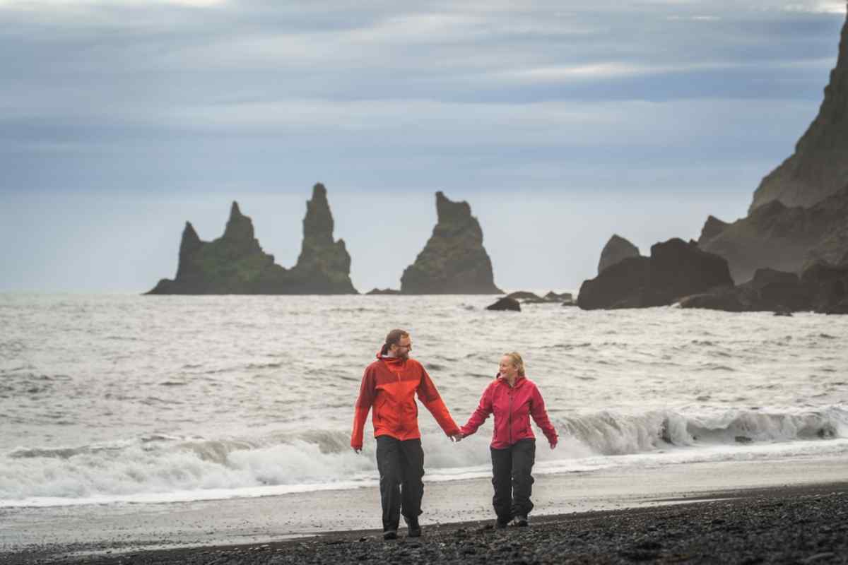 Pareja con chaquetas rojas camina por la playa de Reynisfjara, arena negra y olas, con los farallones de Reynisdrangar al fondo en día nublado.