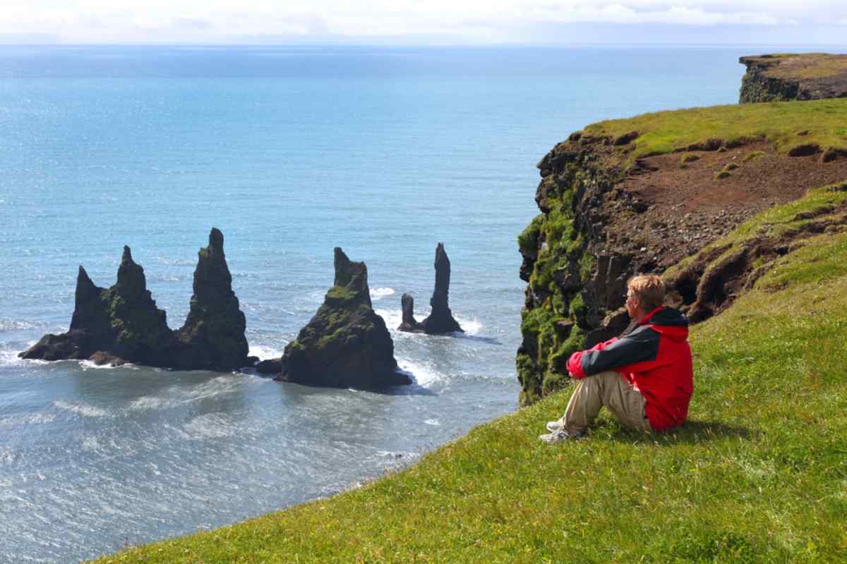 Viajero con chaqueta roja sentado en un acantilado herboso frente a los farallones de Reynisdrangar, con vista de la costa sur de Islandia.