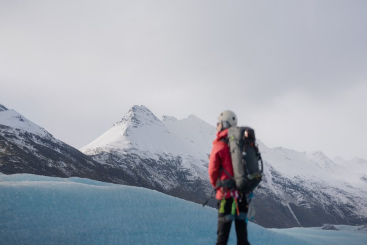 Senderista sobre glaciar con chaqueta roja junto a hielo azul y montañas nevadas islandesas al fondo